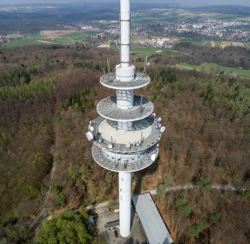 Luftaufnahmen und Drohnenfotografie: Ein Funkturm im Wald. Im Hintergrund sieht man den nächst gelegenen Ort mit Wiesen und Straßen.