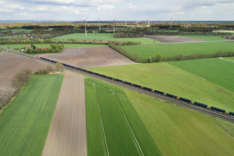 Luftaufnahmen und Drohnenfotografie: Ein Güterzug transportiert Pipelinerohre quer durch Deutschland. Das Gleis führt durch grüne Wiesen und Felder. Im Hintergrund sieht man Windkraftanlagen.