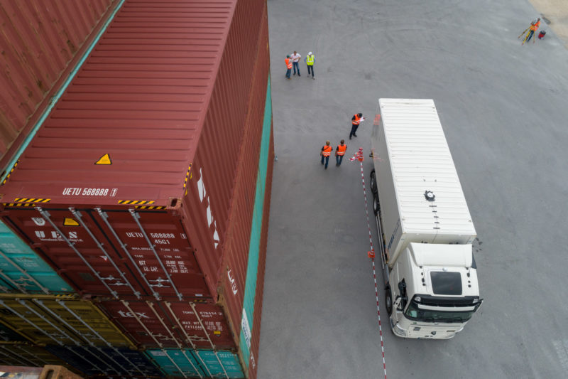 Luftaufnahmen und Drohnenfotografie: Mehrere Mitarbeiter in Schutzwesten arbeiten am Test eines LKW mit Positionierungssystem in einem Containerlager. 