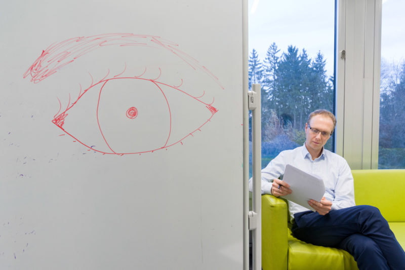 Editorial Fotografie: Wissenschaftler forscht an der Schnittstelle zwischen Mensch und Maschine und der dafür notwendigen visuellen Interaktion. Portrait mit Tablet auf dem Sofa und einem gezeichneten Auge an der Wand.