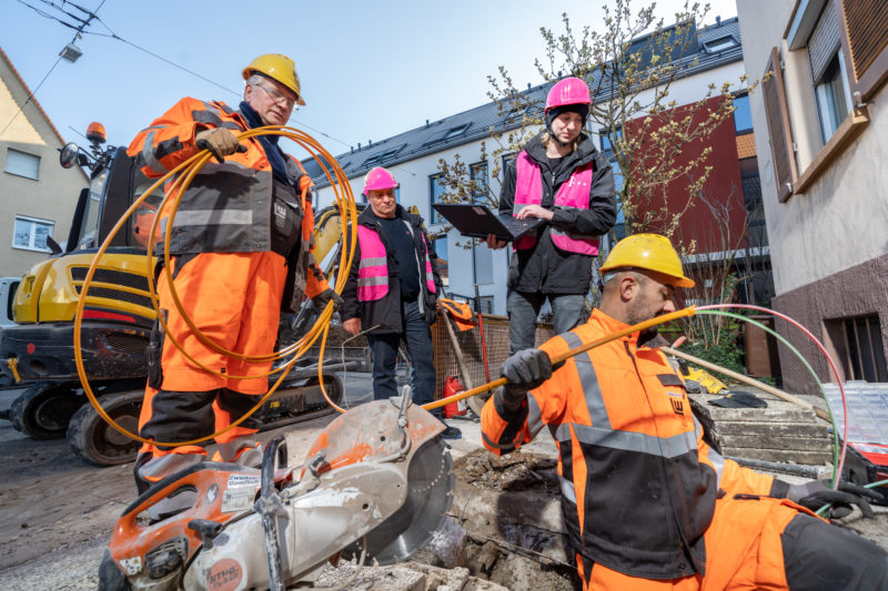 Reportagefotografie auf einer Glasfaserbaustelle: Die Mitarbeiter der Telekommunikationsfirma arbeiten mit den Arbeitern der Baufirma eng zusammen, damit die Glasfaserleitungen für schnelles Internet korrekt verlegt werden.