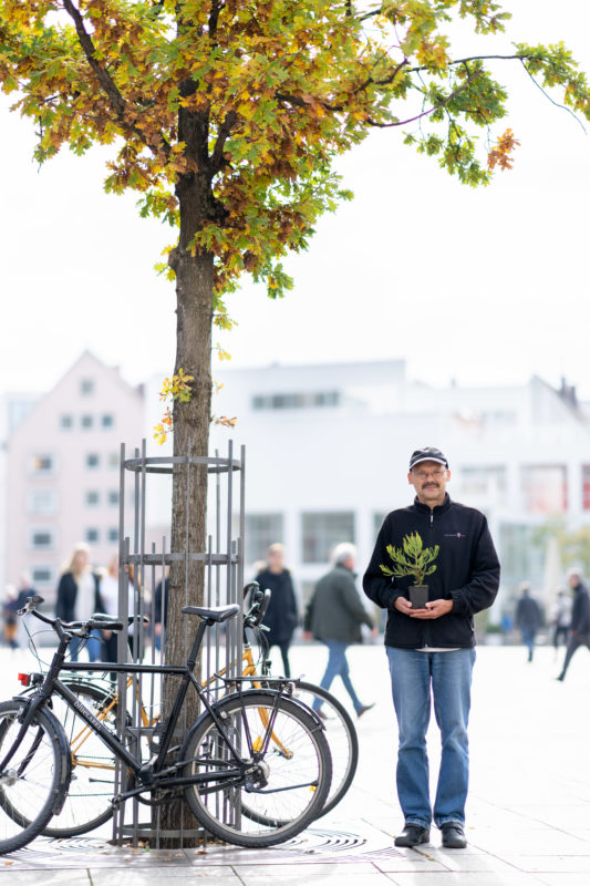 Editorial Portrait: Ein Mitarbeiter der sich für das Pflanzen von Bäumen einsetzt. Er steht im Gegenlicht auf einem belebten Platz unter einem Baum und hat einen Setzling in der Hand.