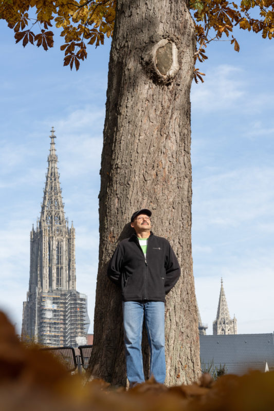 Editorial Portrait: Ein Baumfreund lehnt an einem alten Baum und schaut in die Sonne. Dahinter die Türme einer großen Kirche.