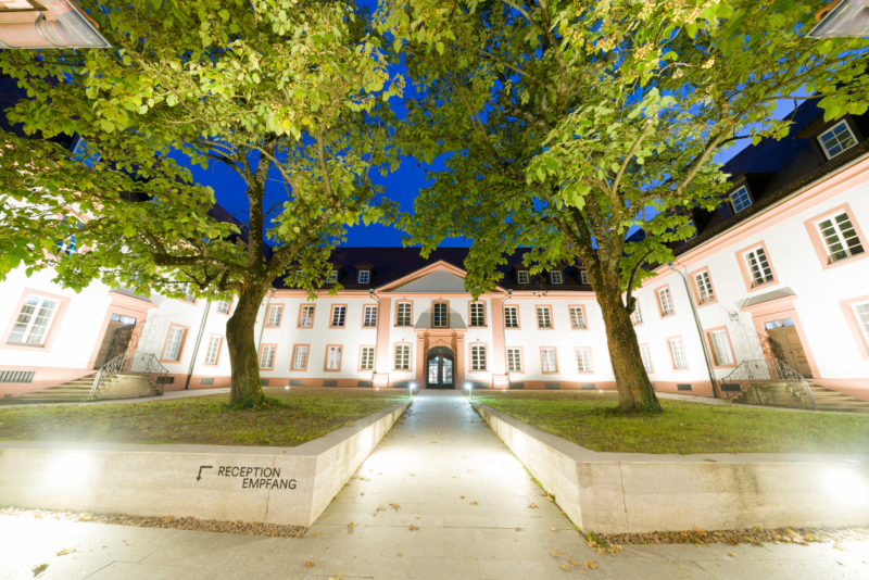 Architekturfotografie: Der nächtlich beleuchtete Innenhof eines ehemaligen Klosters, das nun ein privates College beherbergt. Bäume und Grünflächen säumen den Weg zum Haupteingang.