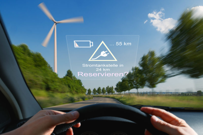 Fotomontage Elektromobilität: Ein Head-Up Display hilft bei Reservieren der nächsten Stromtankstelle. Im Hintergrund ein Windrad.