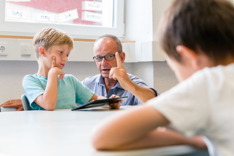 Reportagefotografie: Ein Lehrer bei der Arbeit: Reportagefotografie: Ein Lehrer für Englisch erklärt einem seiner Schüler etwas. Dabei bittet er mit dem Zeigefinger um Aufmerksamkeit und der Schüler hört ihm genau zu.