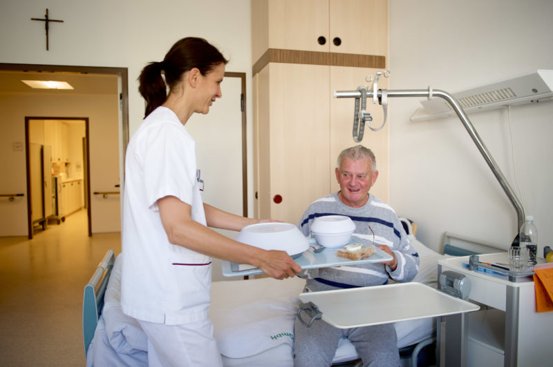 Medizinfotografie: Eine Pflegekraft bringt das mit Isolierhauben versehene Essen auf einem Tablett ans Bett eines Patienten eines Kreiskrankenhauses. Er sitzt bereits wartend auf der Bettkante.