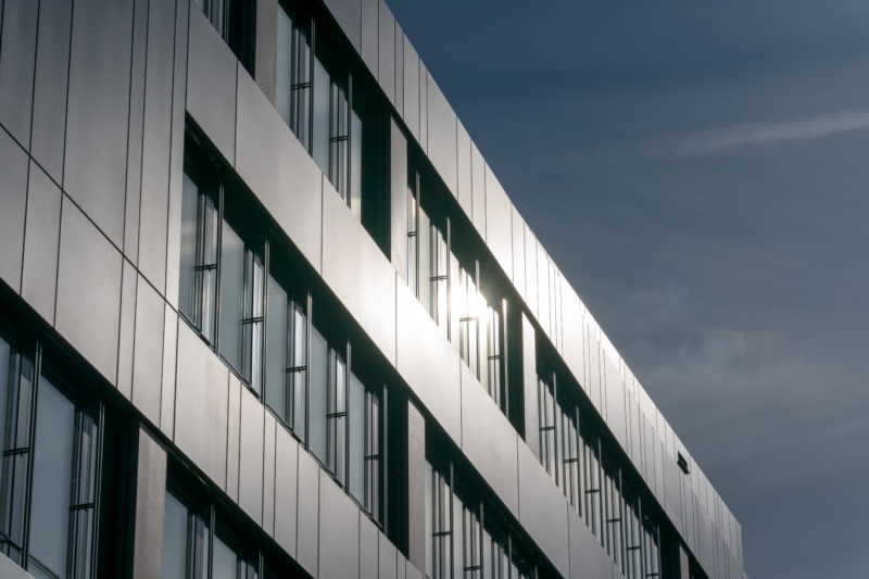 Architekturfotografie: Mit Teleobjektiv von unten nach oben fotografierte Außenansicht der dunklen Fassadenelemente aus Metall eines neuen Bürogebäudes. Im Metall spiegelt sich die Sonne, dahinter sieht man den mit leichten Wolken bedeckten Himmel.