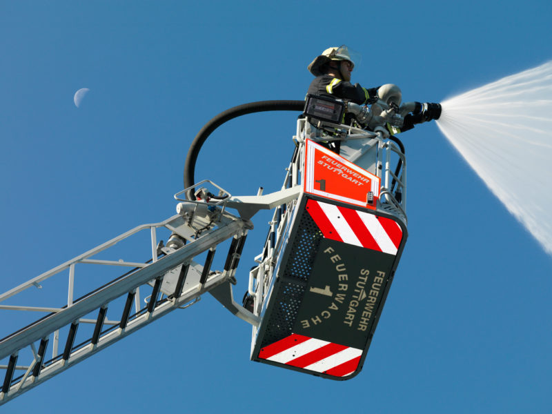 Reportagefotografie: Ein Feuerwehrmann lenkt im Korb einer Drehleiter das geöffnete Strahlrohr. Am blauen Himmel dahinter steht die Sichel des abnehmenden Mondes.