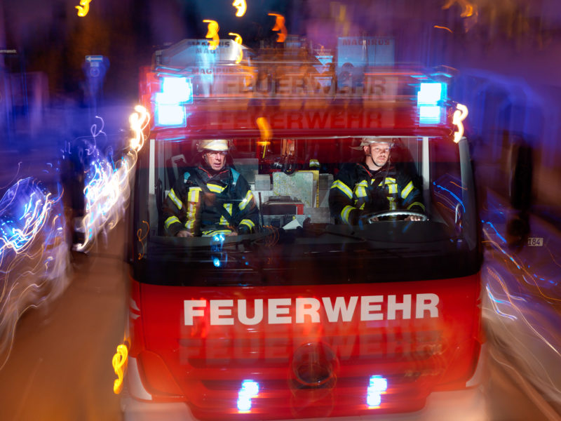 Luftaufnahmen und Drohnenfotografie: Reportagefotografie: Zwei Feuerwehrleute in ihrem Hilfeleistungslöschfahrzeug auf der nächtlichen Fahrt mit Blaulicht.