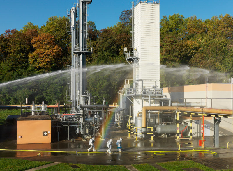 Reportagefotografie: Bei einer Übung an einem Gasverteiler gehen drei Feuerwehrleute in silberner Hitzeschutzkleidung und Werkzeug nach vorne, während über ihnen ein Löschmonitor Wasser zum angenommenen Brandherd bringt.