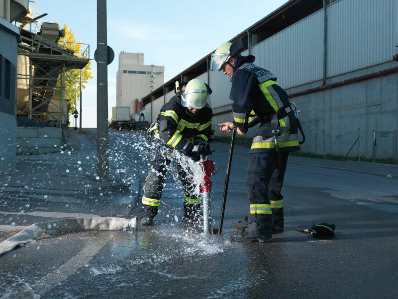 editorial photography: Two firemen attach a standpipe to a hydrant in the ground, to which fire hoses can then be connected. Water splashes.