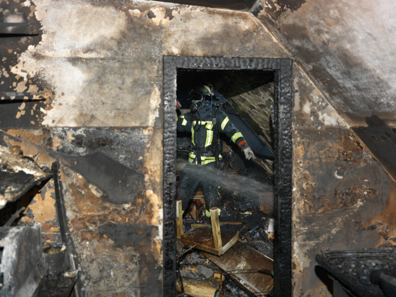 Reportagefotografie: Feuerwehrmann beim Nachlöschen in einem ausgebrannten Dachstock eines Wohnhauses. Alles ist schwarz verkohlt und auch das ganze Mobiliar ist zerstört.