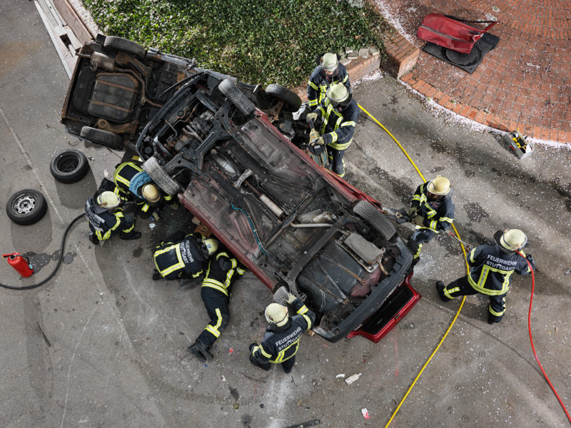 Reportagefotografie: Feuerwehrleute üben die Bergung von eigeklemmten verletzten Insassen eines verunglückten PKW. Das Foto ist senkrecht von oben aufgenommen und man sieht in der Mitte den Boden des auf dem Dach liegenden Fahrzeuges, das von Feuerwehrleuten umringt ist.