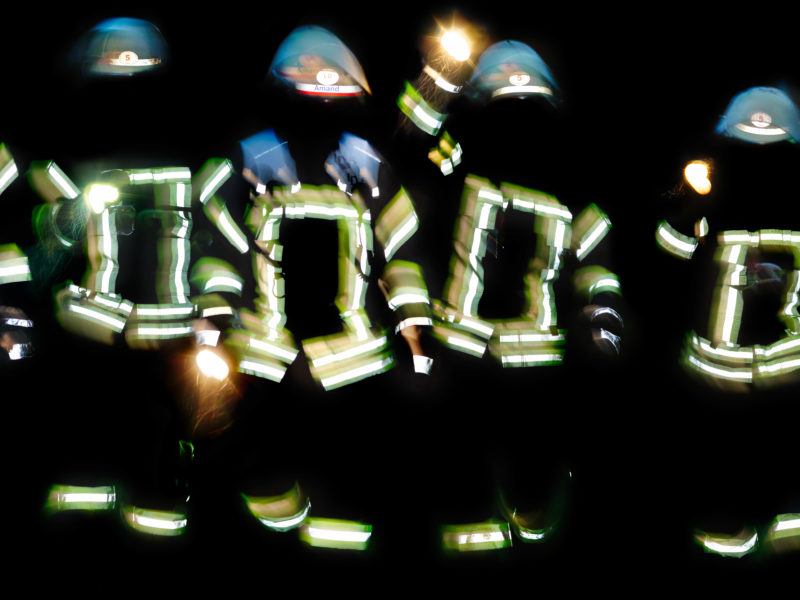 editorial photography: Firemen at night. The signal stripes on the jackets and helmets reflect brightly while the faces remain in the dark.