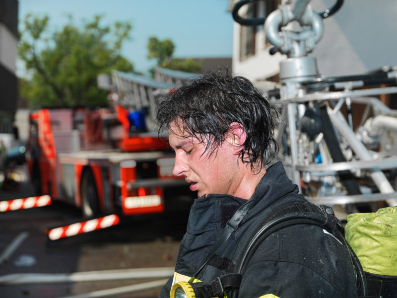 Reportagefotografie: Ein Feuerwehrmann, nachdem er bei seinem Löscheinsatz in einem brennenden Haus abgelöst wurde. Schweiss bedeckt seinen Kopf.