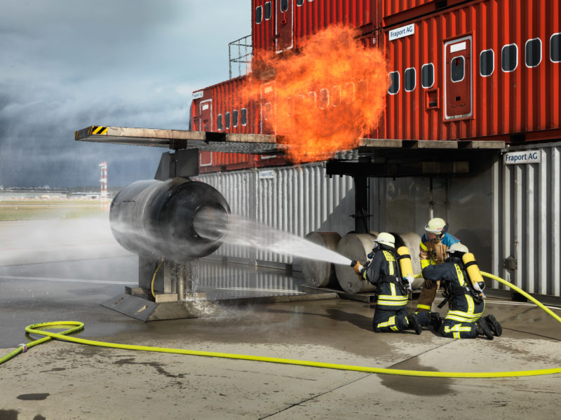 Reportagefotografie: Bei einer Großübung am Flughafen spritzen Feuerwehrleute Wasser gezielt in die Attrappe einer Flugzeugturbine, nachdem von dort eine Flammenexplosion aufstieg.