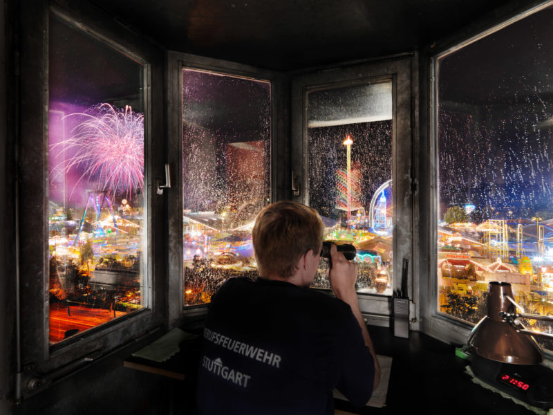 Reportagefotografie: Ein Stuttgarter Berufsfeuerwehrmann überwacht nachts aus einer Kanzel in der benachbarten Feuerwache das Cannstatter Volksfest, während dort das Feuerwerk gezündet wird.