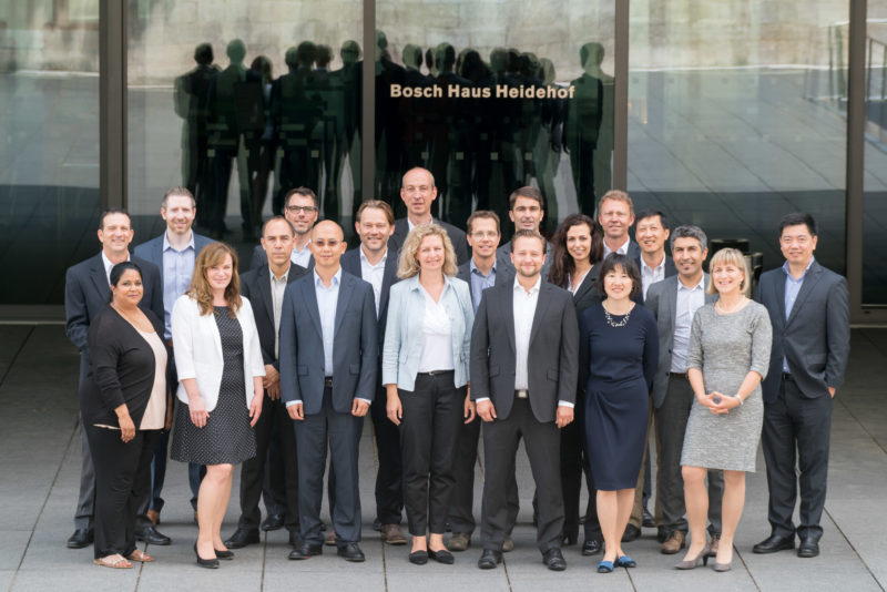 Group photo: The members of a department stand together in front of a company building. They are reflected as silhouettes in the glass panes behind them.