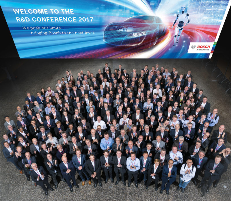 Group photo: About 200 employees of a company conference at a group picture in the evening. The photo is illuminated with flash and a large banner with the conference title hangs over the heads of the participants.