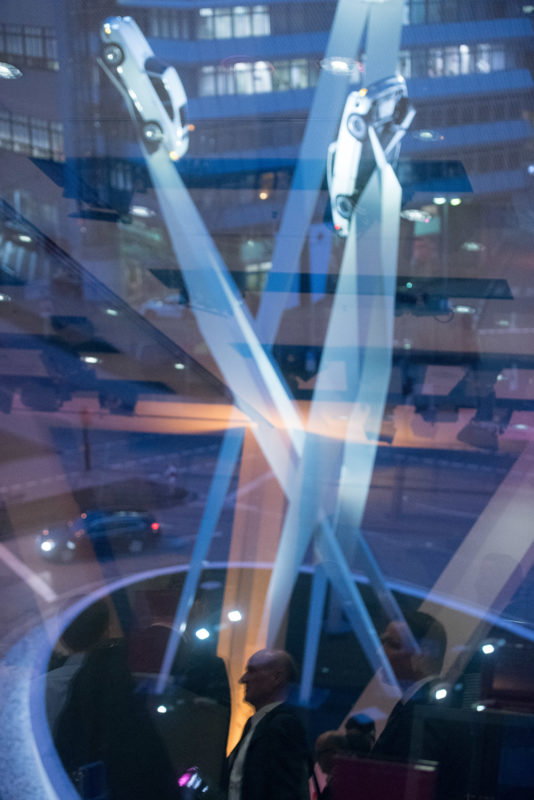 Editorial photography as event photography and fair photography: At a company event at the Porsche Museum in Stuttgart, participants are reflected in the large windows, while outside you can see the work of art illuminated at night on the roundabout in front of the museum, on which Porsche cars symbolically drive into the sky.