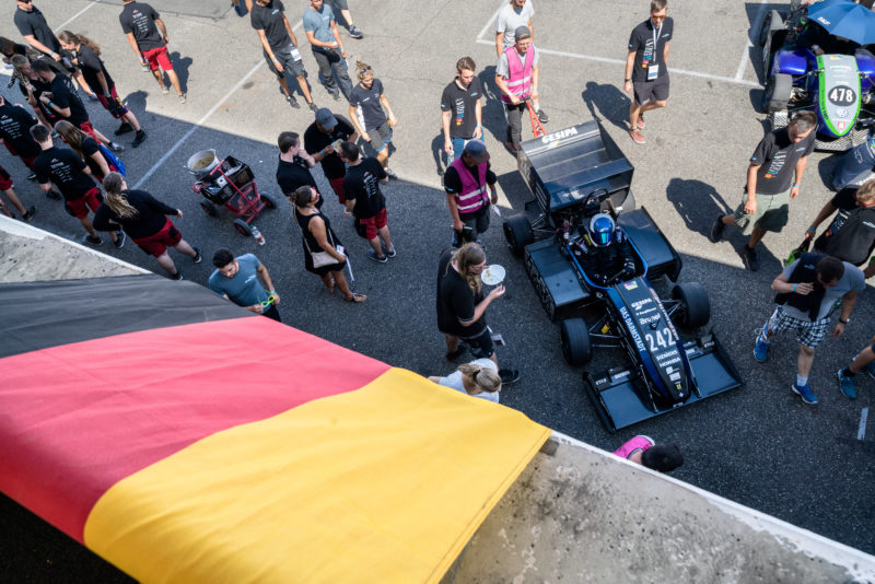 Reportagefotografie: Formula Student Germany: Ein Rennwagen wird gemeinsam aus der Sonne in die Box gebracht. Über dem Tor hängt die deutsche Fahne.