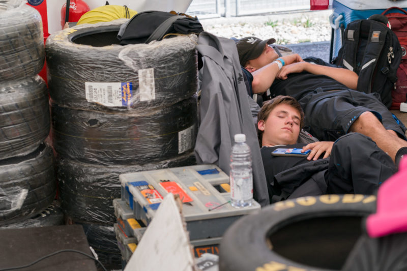 Reportagefotografie: Formula Student Germany: Zwei Studenten liegen in einer Rennpause in der Box schlafend zwischen gestapelten Reifen und Material.