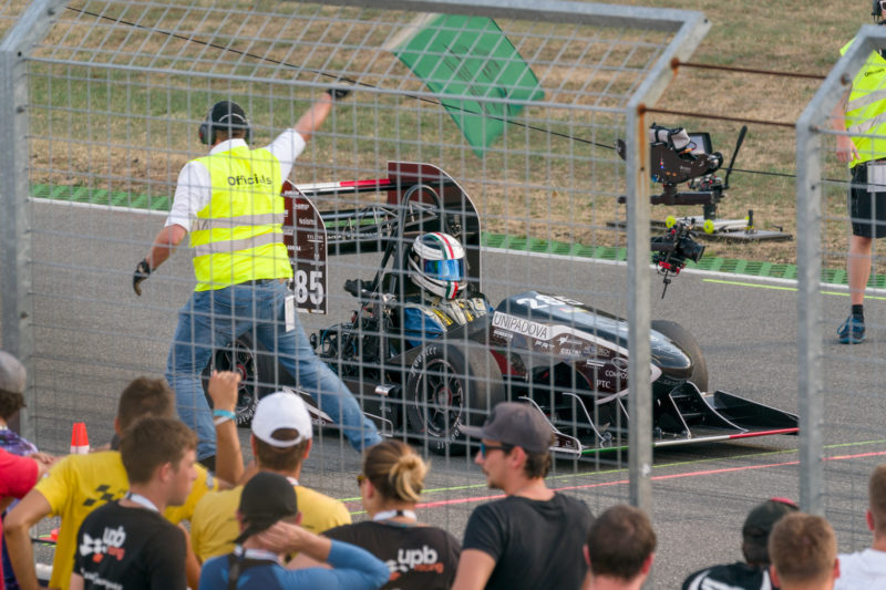 Reportagefotografie: Formula Student Germany: An der Rennstrecke zur Beschleuningsmessung startet ein Rennwagen seinen Lauf. Zuschauer sind im Vordergrund und eine Seilkamera soll sein Rennen begleiten.