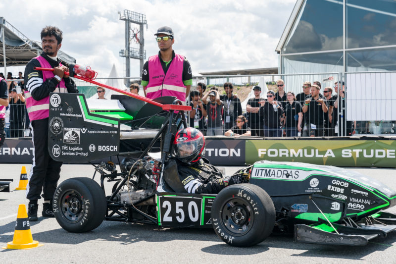 Reportagefotografie: Formula Student Germany: Teammitglieder aller Teams schauen von der Zuschauerseite aus auf die im Rennen fahrenden Wagen. Vorne wartet ein Verbrennerrennwagen auf seinen Start.