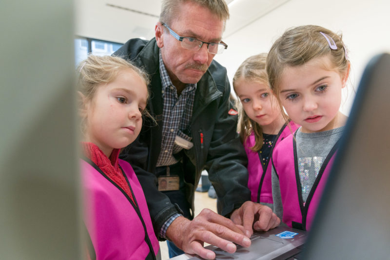 Reportagefotografie zum Thema Lernen und Bildung: Drei kleine Mädchen schauen in einem Betrieb an einem Tüftler- und Forscherinnentag einem Techniker zu, wie er am Notebook an der Steuerung einer Maschine arbeitet.