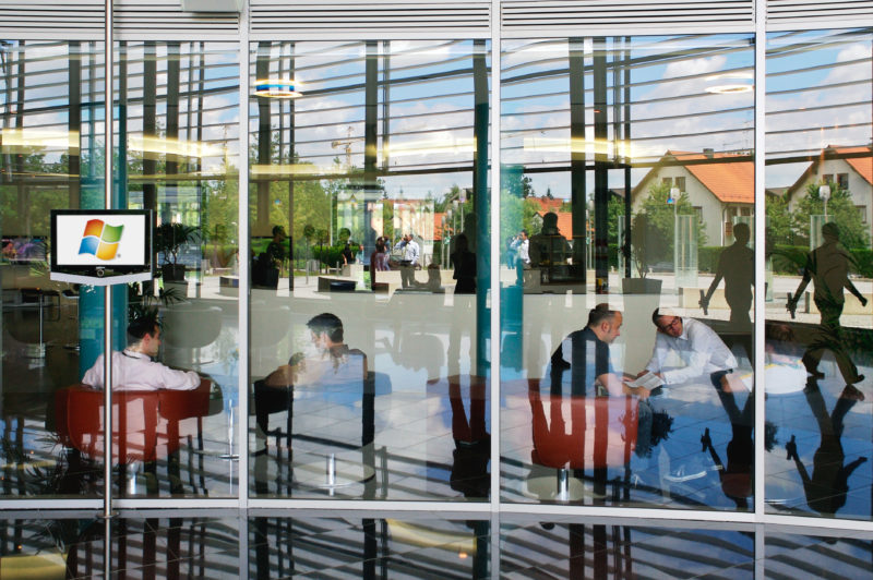 Architectural photography:  In the entrance area of a modern office building, the people in the foyer and in a lounge merge with the architecture of the glass facade and the buildings in the neighbourhood that are reflected in it.