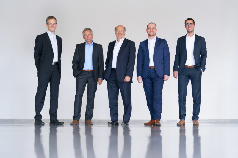 Group photo: The 5 executives of a company in full body portrait in front of a light neutral background and on a light reflecting floor.