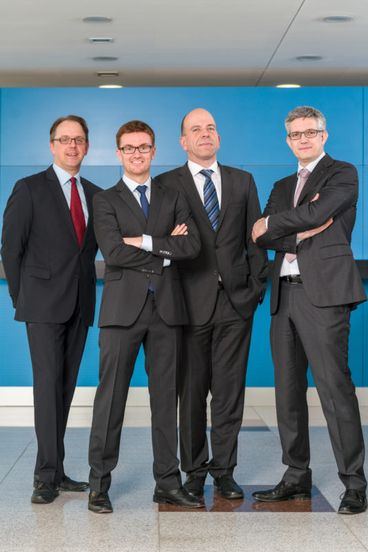 Group photo:  Four executives in the foyer of their company. Full body group picture with suit and tie.