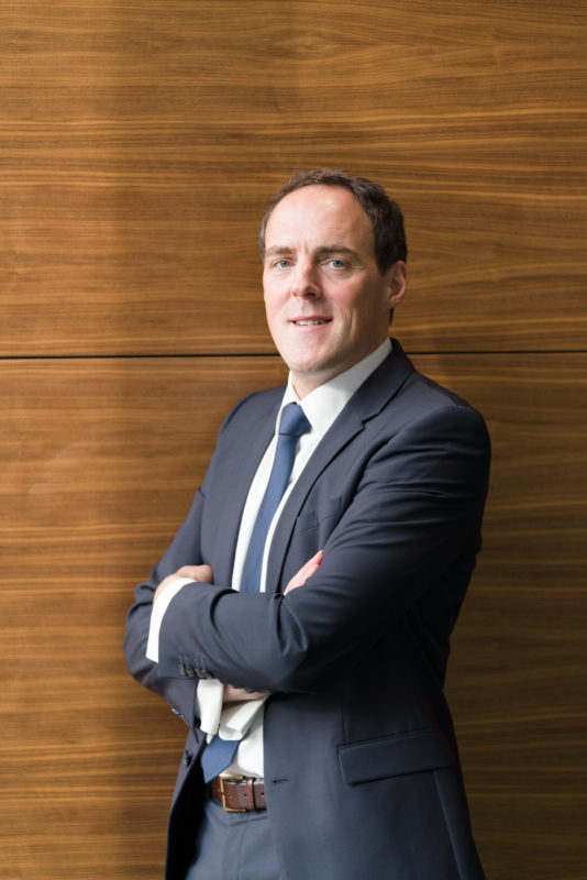 Managerportrait: Male leader in half portrait in front of a brown cross-striped wooden wall. He has crossed his arms and is illuminated by a flashgun with softbox half from the front left. The focal length used is in the middle telephoto range.