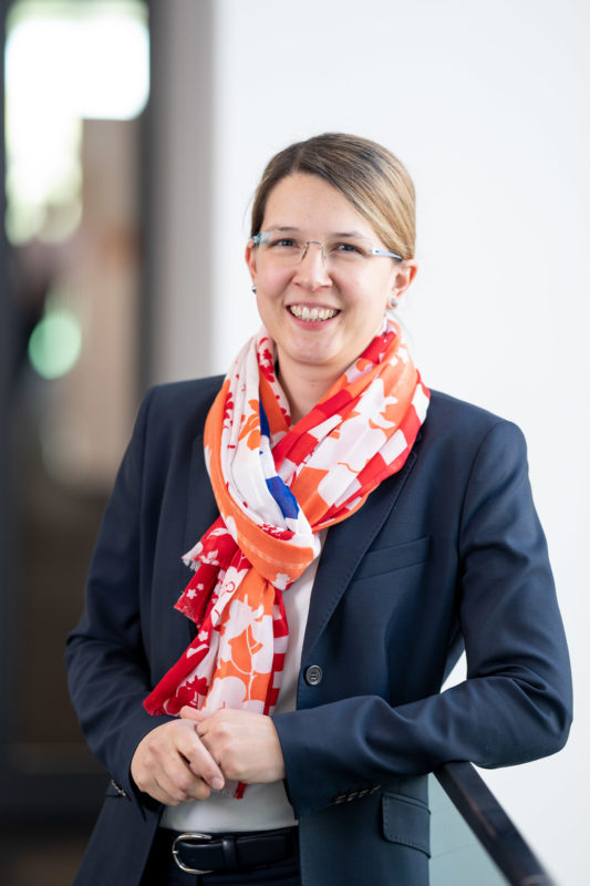 Executive portrait: A young woman with glasses and coloured scarf laughs into the camera. In the background you can see beautiful blurred company surroundings.