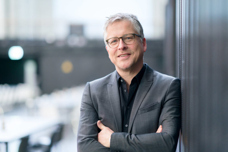 Managerportrait: Executive in black shirt and jacket. He leans against a dark metal wall. The background is deliberately blurred, the sharpness lies on the eyes of the man.