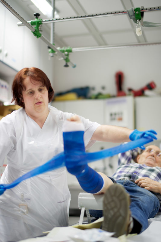 Healthcare photography:  In the plaster room a nurse cares for the foot of an elderly male patient. She uses a blue plastic bandage, which hardens later and thus stabilizes the fracture.