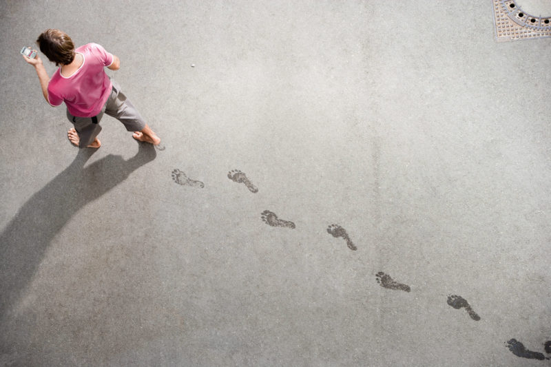 Special perspective from above: A man walks across a street with his smartphone and leaves marks on the asphalt with his wet feet