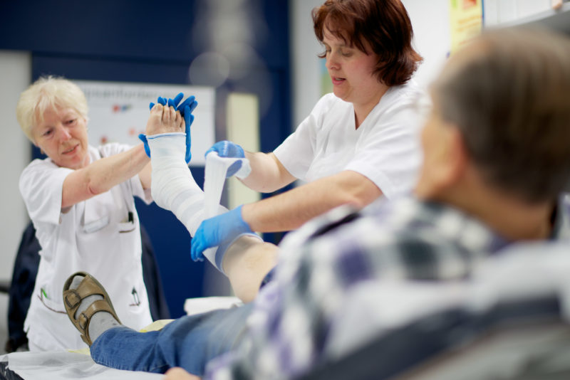 Medizinfotografie: Versorgung eines älteren Patienten im Gipsraum. Zwei Pflegerinnen wickeln einen Verband um seinen rechten Fuss.