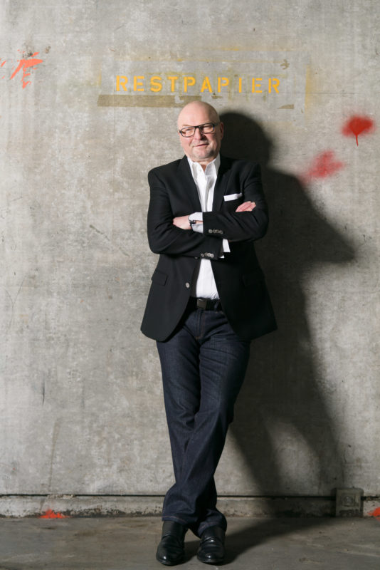 Managerportrait: Full body portrait of a boss of a publishing printing house. With his arms crossed, he leans with his back against a concrete wall in the warehouse of his printing company. The hard light of the flash casts a shadow on the wall. Above him the word Restpapier stands on the wall and you can see spots of paint on the concrete.