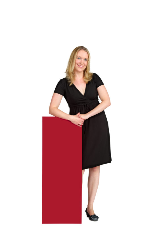 Employees photography: For a personnel campaign of a vehicle manufacturer, employees pose with a red sign against a white background. The photos are full body shots and each person acts differently in front of the camera. Here a woman just stands there relaxed.