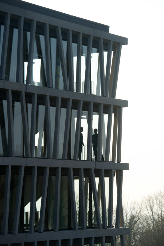 Architekturfotografie: Grafisch wirkende Außenaufnahme eines Gebäudeteils eines modernen Forschungsinstitutes. Durch die offene Fassadenstruktur sieht man die Silhouette von Personen im Gebäude, die sich unterhalten.