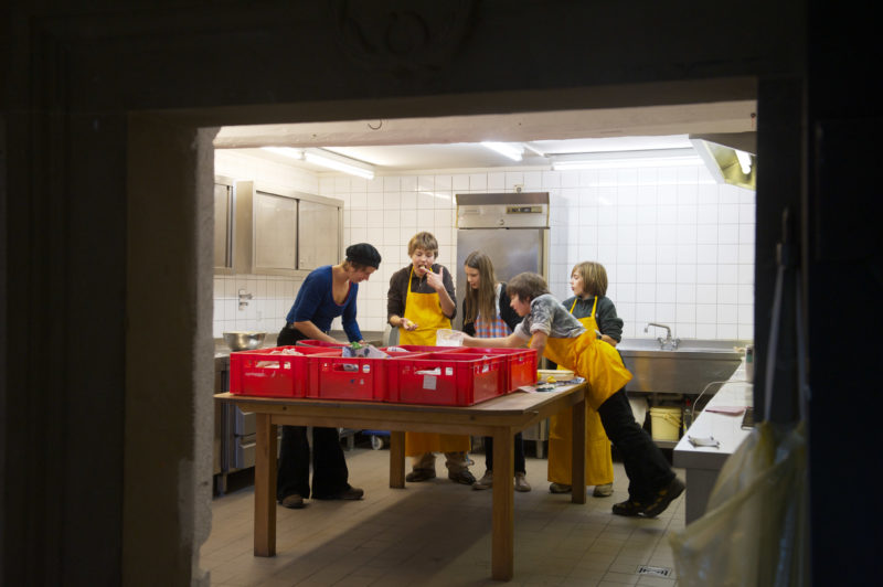 Reportagefotografie: Teil einer Schulklasse beim gemeinsamen Kochen in der Schulküche. Alle tragen Schürzen und einer der Jungen probiert gerade einen großen KlumpenTeig.