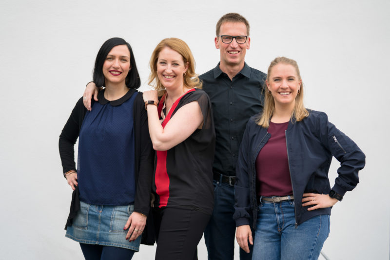 Group photo: Portrait of four speakers of an internal company event series, taken a few minutes before the start of the event in natural light and against a calm, bright background.