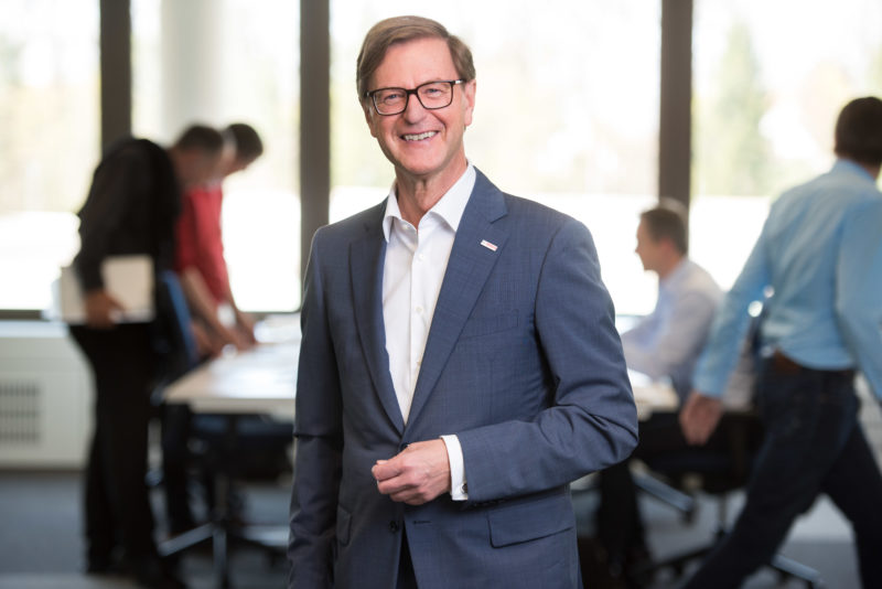 Managerportrait: A managing director stands in the office and laughs into the camera. It is illuminated with a flash, while in the background employees are still recognizable at tables.