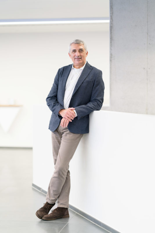 Managerportrait:  An Italian businessman in a blue jacket and beige trousers leans casually against a balustrade. The large room in the company looks bright and friendly.