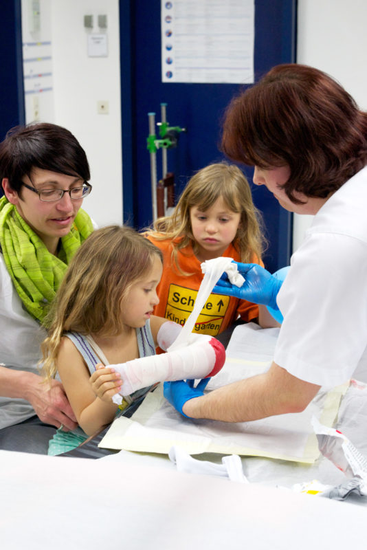 Healthcare photography: A nurse wraps the broken arm of a little girl in a cast. Her little sister and her mother are with her.