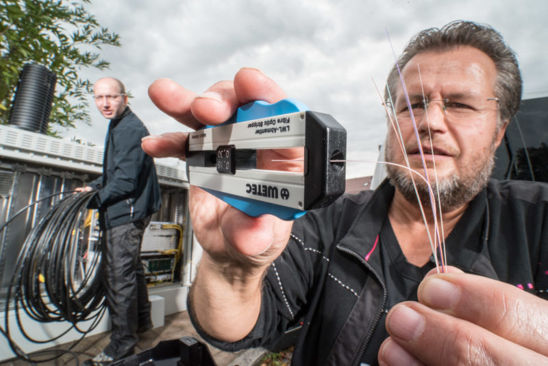 Reportageotografie: Ein Techniker mantelt auf der Straße ein Glasfaserkabel ab, während ein Kollege im Hintergrund am Anschluss eines Verteilergehäuses für Breitbandinternetverbindungen arbeitet.