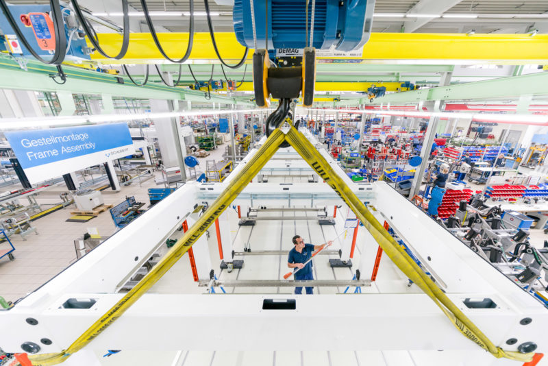 Industriefotografie: Ein Mitarbeiter eines Herstellers für Verpackungsanlagen arbeitet am Grundgestell einer Maschine. Auf dem Weitwinkelfoto sieht man weiteren Arbeitsplätze in der großen Montagehalle.