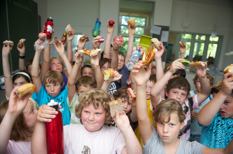 Reportagefotografie: Kinder einer Grundschulklasse halten die mitgebrachten Speisen in die Kamera, die sie in ihrer Pause gemeinsam essen. Trinken dürfen sie zu jeder Zeit des Unterrichts.